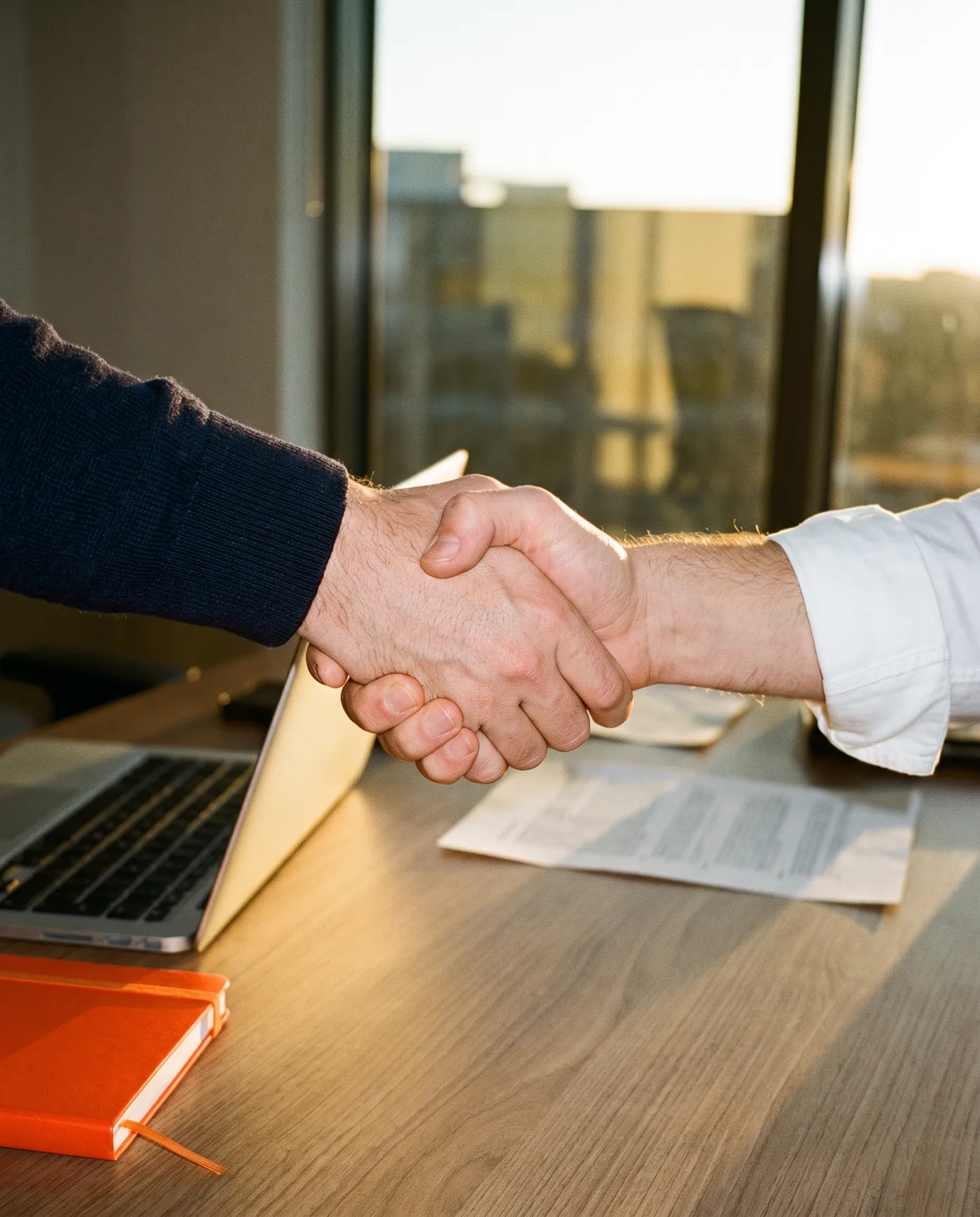 Two hands in a firm handshake over a desk in golden evening light, a laptop and an orange notebook beside them